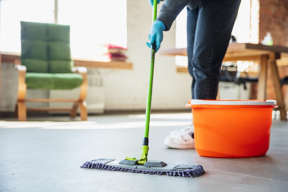 Person cleaning floor with a mop next to an orange bucket in a bright room.
